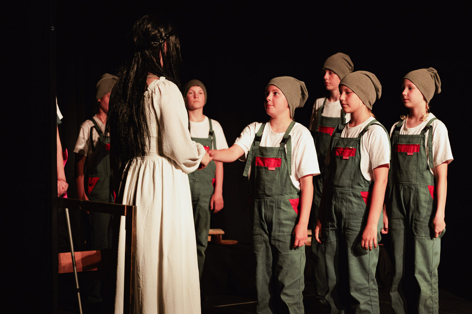 Auf einer dunklen Bühne steht eine Frau in einem weißen Kleid mit langen schwarzen Haaren einer Gruppe von Kindern gegenüber. Die Kinder tragen grüne Latzhosen, weiße T-Shirts und graue Mützen, während eines von ihnen der Frau die Hand gibt.