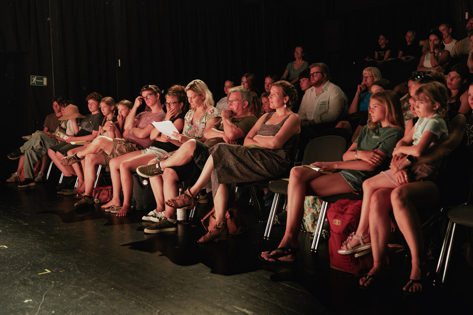Ein Publikum sitzt in einem dunklen Theaterraum auf Stühlen und schaut aufmerksam nach vorne zur Bühne. Warmes Licht beleuchtet die Gesichter der Erwachsenen und Kinder, während der Hintergrund fast vollständig schwarz bleibt.