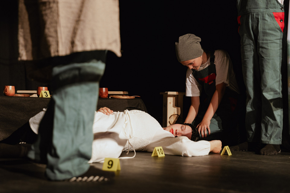 Auf einer dunklen Bühne liegt eine Person in weißer Kleidung am Boden, umgeben von gelben Markierungsschildern wie an einem Tatort. Mehrere Personen in Arbeitskleidung stehen oder knien daneben, wobei eine Person den Kopf der liegenden Person berührt.