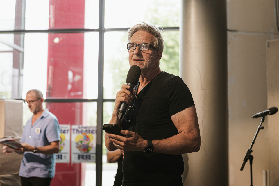 Ein Mann mit Brille hält ein Mikrofon und ein Smartphone in der Hand, während er in einem hellen Innenraum spricht. Im Hintergrund steht ein weiterer Mann mit Unterlagen, daneben sind große Fenster, eine Säule und ein Mikrofonständer zu sehen.