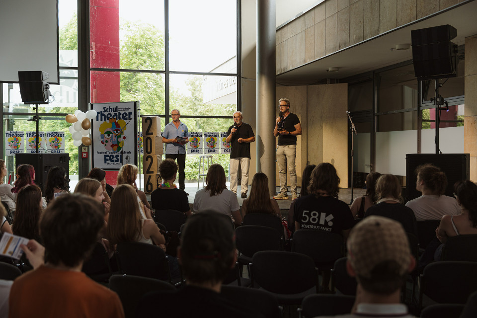 Drei Männer stehen mit Mikrofonen vor einem sitzenden Publikum in einer modernen Halle und moderieren offenbar eine Veranstaltung. Neben ihnen sind Plakate und Dekorationen zum Schul-Theater-Fest 2026 zu sehen.