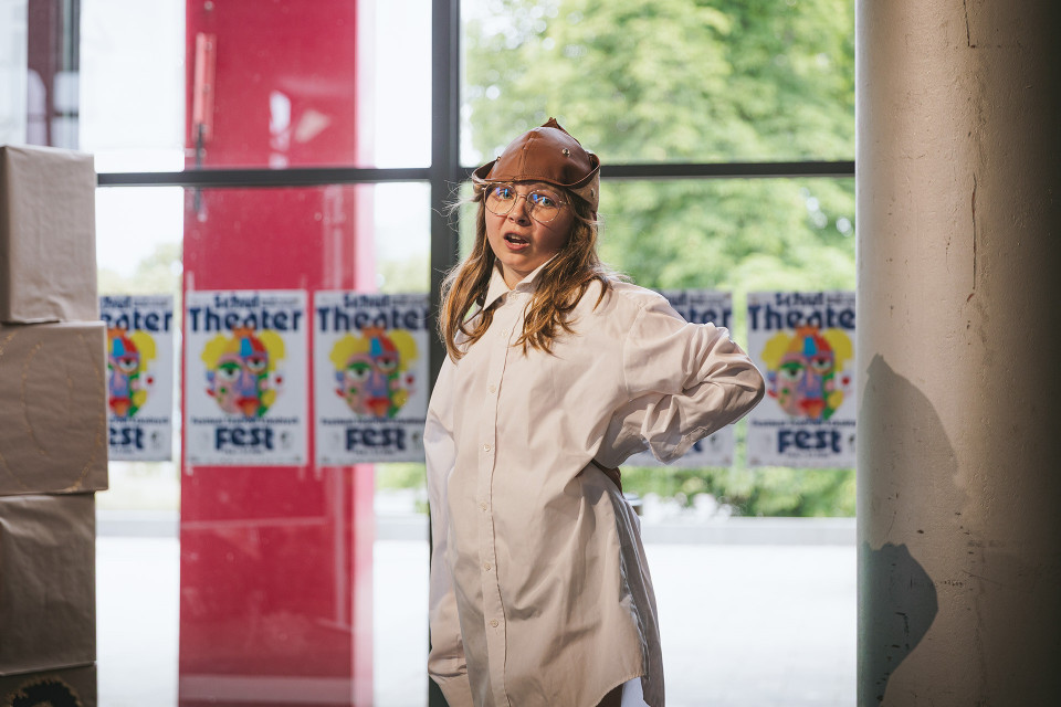 Ein Kind mit Brille, weißem Hemd und brauner Lederkappe steht in einem hellen Innenraum vor großen Fenstern. Im Hintergrund hängen mehrere Plakate für ein Theaterfest, während draußen grüne Bäume zu sehen sind.