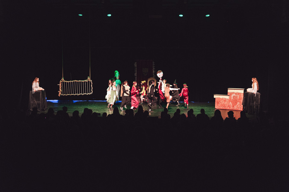 Auf einer dunklen Theaterbühne führen mehrere Kinder in bunten Kostümen eine Szene auf, während links eine hängende Requisite und rechts ein bemaltes Bühnenelement zu sehen sind. Im Vordergrund sitzen die Zuschauer als dunkle Silhouetten, während die Bühne von wenigen grünen Scheinwerfern beleuchtet wird.