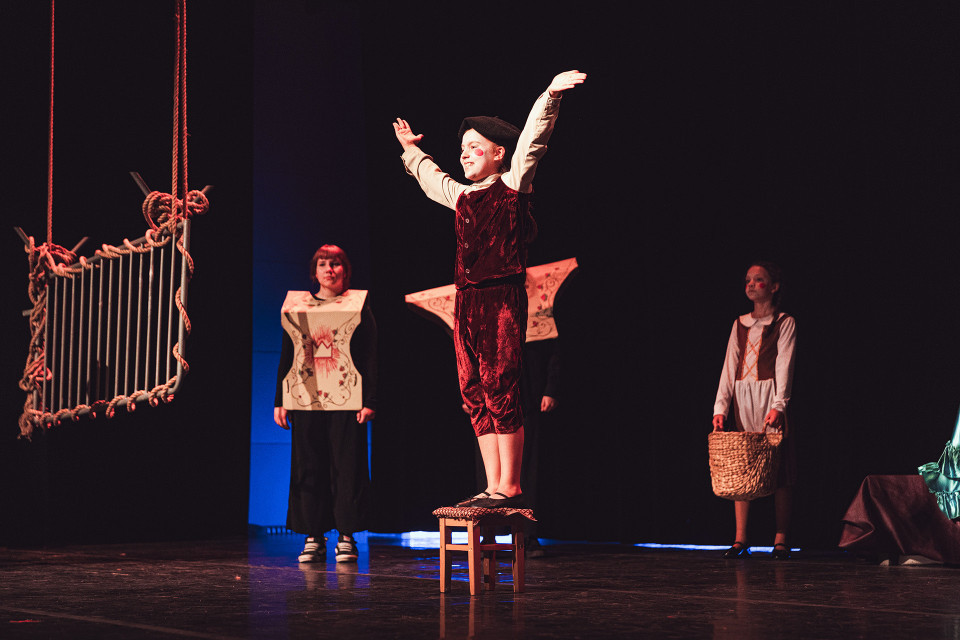 Auf einer dunklen Theaterbühne steht eine junge Darstellerin in einem roten Kostüm auf einem Hocker und hebt die Arme. Um sie herum stehen weitere kostümierte Personen, während links eine hängende Requisite aus Seilen zu sehen ist.