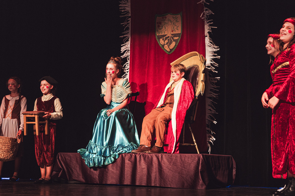 Kinder führen auf einer Bühne ein Theaterstück in historischen Kostümen auf, mit einer Prinzessin im türkisfarbenen Kleid und einem Jungen im roten Umhang auf einem erhöhten Podest. Im Hintergrund hängt ein roter Vorhang mit Wappen, während weitere Kinder mit roten Wangen geschminkt am Rand der Szene stehen.