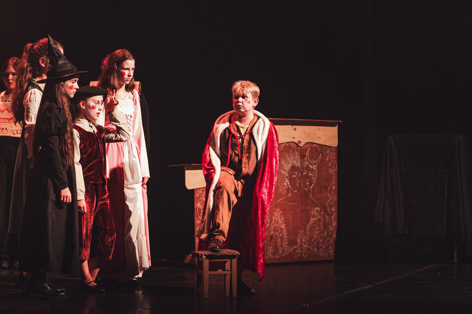 Auf einer dunklen Theaterbühne stehen mehrere Kinder in fantasievollen Kostümen, darunter eine Hexe, ein Junge in rotem Samt und eine Figur in einem weißen Kleid. Rechts sitzt ein blondes Kind mit rotem Umhang auf einem Stuhl vor einer bemalten Kulisse und blickt zu der Gruppe hinüber.