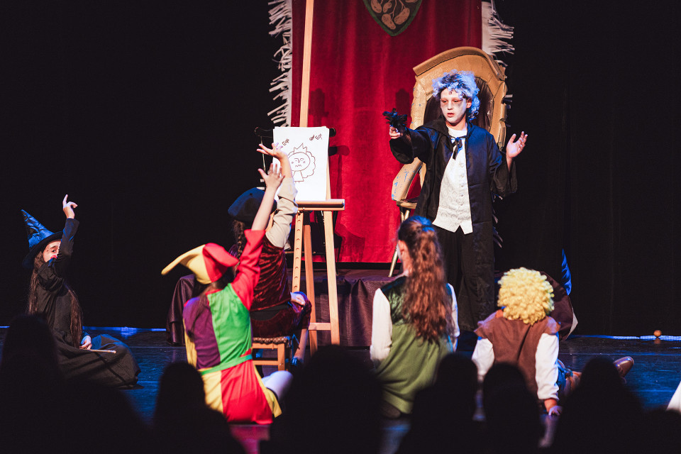 Kinder führen auf einer Bühne ein Theaterstück auf, verkleidet als Zauberer, Narr und andere Figuren. Ein Kind mit blauer Perücke steht vor einem großen roten Vorhang neben einer Staffelei, während die anderen Kinder zu ihm hinaufschauen und die Hände heben.
