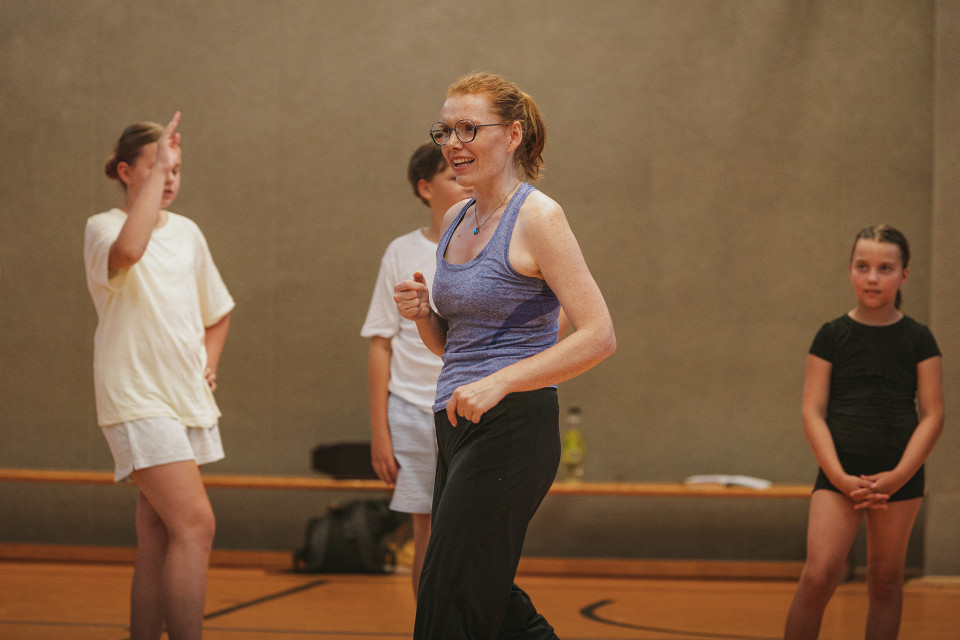 Eine lächelnde Frau in Sportkleidung steht in einer Turnhalle und scheint eine Übung oder Tanzbewegung anzuleiten. Um sie herum stehen mehrere Kinder in Sportkleidung, die aufmerksam mitmachen oder zuschauen.