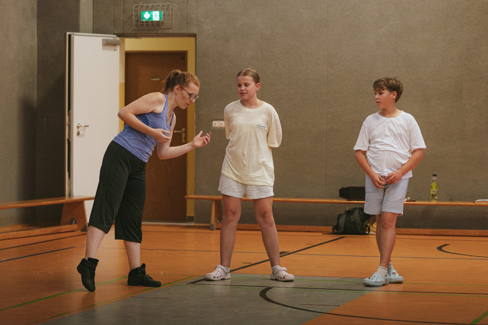 Eine Frau steht in einer Sporthalle und gestikuliert, während sie mit zwei Kindern spricht. Die Kinder tragen helle Sportkleidung und stehen auf dem Hallenboden vor einer grauen Wand und einer offenen Tür.