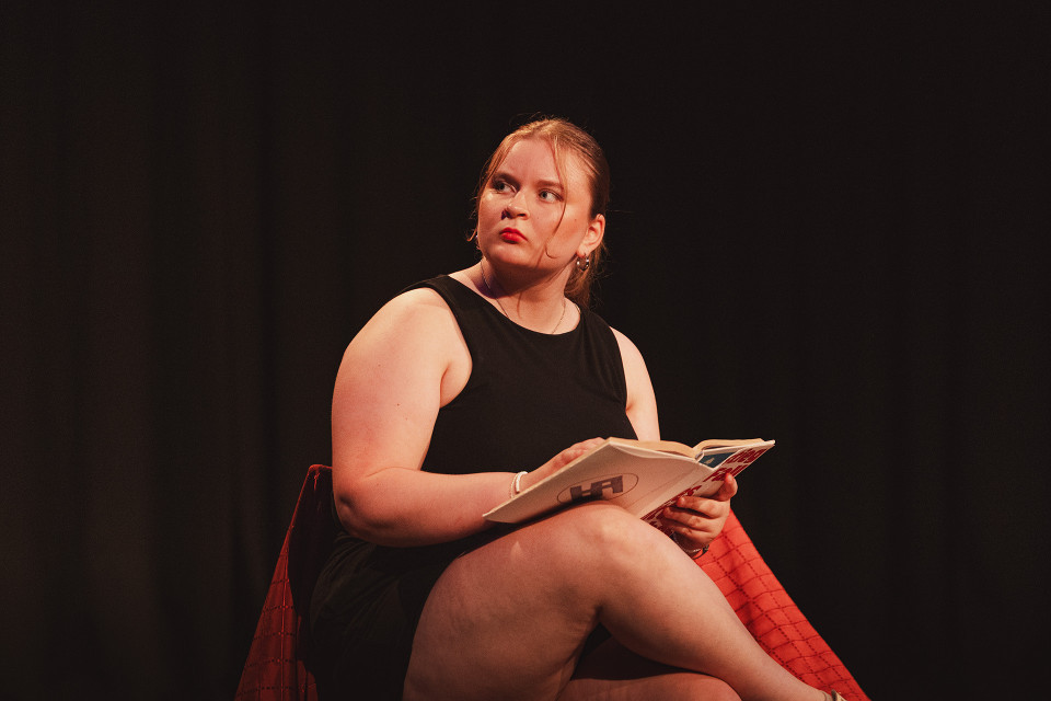 Eine junge Frau in einem schwarzen ärmellosen Kleid sitzt auf einem roten Stuhl und hält ein aufgeschlagenes Buch in den Händen. Vor dunklem Hintergrund wird sie warm beleuchtet und blickt mit ernstem Ausdruck zur Seite.