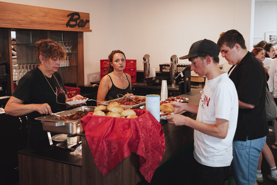 An einer Bar werden mehrere Personen mit warmem Essen wie Würstchen und Brötchen bedient. Hinter der Theke stehen Mitarbeiterinnen, während Gäste mit Tellern in einer Schlange warten.