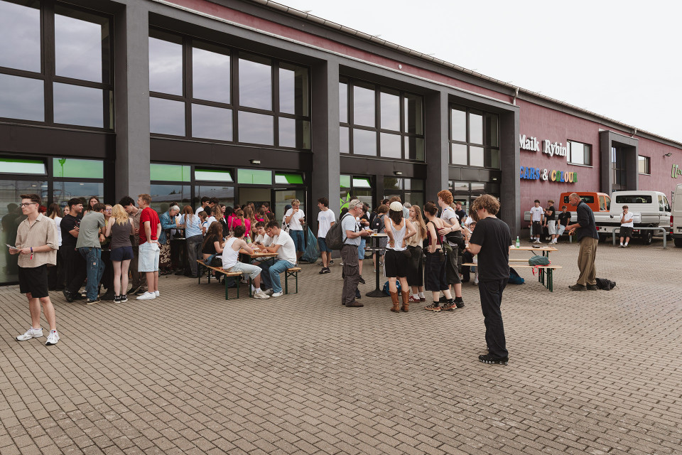 Vor einem modernen Gewerbegebäude mit großen Fenstern versammelt sich eine größere Gruppe von Menschen auf einem gepflasterten Platz. Einige sitzen an Biertischen, andere stehen in kleinen Gruppen zusammen, während im Hintergrund Lieferwagen und eine farbige Ladenbeschriftung zu sehen sind.