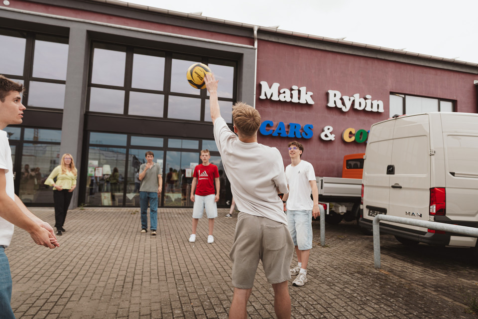 Eine Gruppe junger Menschen spielt auf einem gepflasterten Platz vor einem Gebäude mit der Aufschrift „Maik Rybin Cars & Co“ mit einem gelb-schwarzen Ball. Rechts stehen ein weißer Lieferwagen und ein kleiner Transporter, während mehrere Personen dem Spiel zusehen.