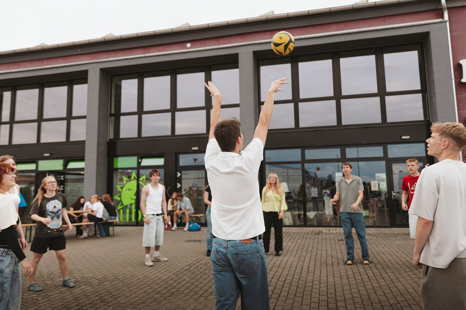 Eine Gruppe junger Menschen spielt auf einem gepflasterten Platz vor einem modernen Gebäude mit großen Fenstern Volleyball. In der Mitte wirft ein Mann in weißem T-Shirt den Ball hoch, während die anderen Spielerinnen und Spieler aufmerksam zusehen.