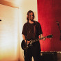 Ein Musiker steht auf einer kleinen Bühne und spielt eine schwarze E-Gitarre vor einem Mikrofon. Warmes Licht, rote Wandverkleidung und weitere Mikrofonständer schaffen eine intime Konzertatmosphäre.
