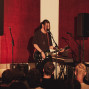 Ein Musiker steht auf einer kleinen Bühne, singt in ein Mikrofon und spielt eine schwarze E-Gitarre. Vor ihm schaut ein Publikum zu, während rote Wandpaneele, Lautsprecher und weiteres Bühnenequipment den Raum prägen.