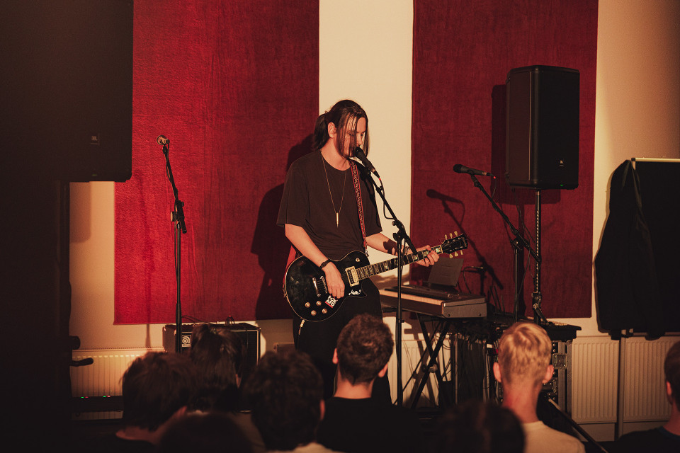 Ein Musiker steht auf einer kleinen Bühne, singt in ein Mikrofon und spielt eine schwarze E-Gitarre. Vor ihm schaut ein Publikum zu, während rote Wandpaneele, Lautsprecher und weiteres Bühnenequipment den Raum prägen.