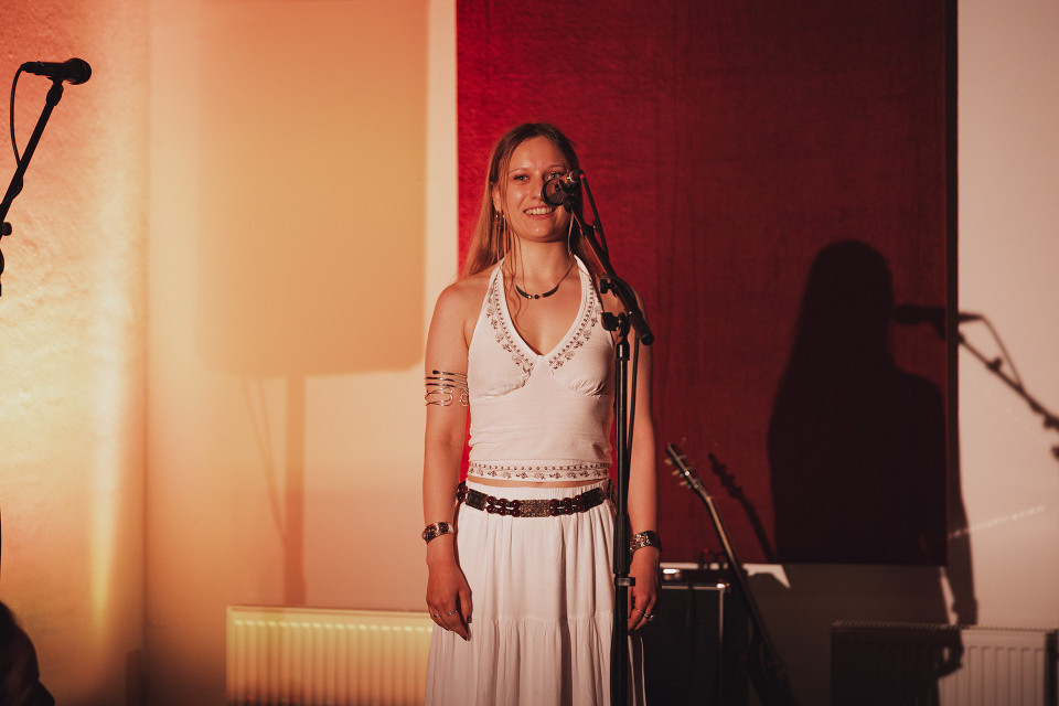 Eine lächelnde junge Frau in einem weißen Outfit steht auf einer Bühne vor einem Mikrofon. Warmes Licht und ein roter Hintergrund werfen ihren Schatten an die Wand, daneben sind weitere Mikrofone und Instrumente zu sehen.