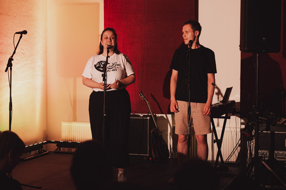 Zwei Personen stehen auf einer kleinen Bühne vor Mikrofonen, während die Frau spricht oder singt und der Mann zu ihr hinüberschaut. Im Hintergrund sind Musikinstrumente, Lautsprecher und warmes Bühnenlicht zu sehen, vor ihnen sitzt ein Publikum im Dunkeln.