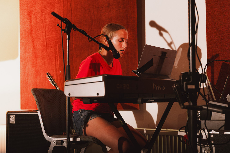 Eine junge Frau sitzt in einem Musikstudio an einem E-Piano und singt in ein Mikrofon. Im Hintergrund sind rote Akustikpaneele, weitere Mikrofonständer und Musikequipment zu sehen.
