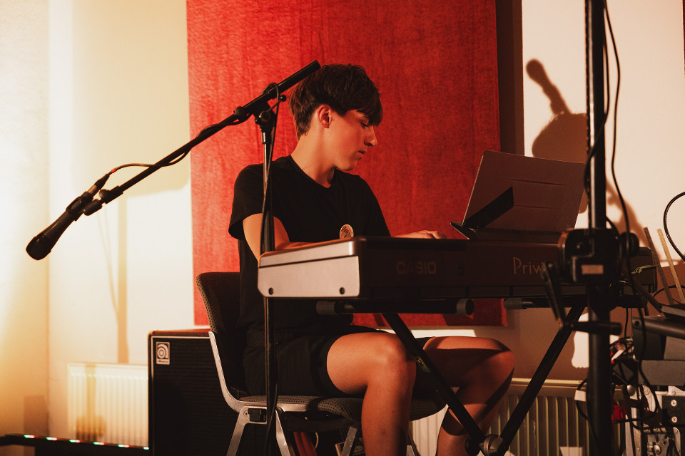 Ein junger Musiker sitzt an einem elektronischen Keyboard und schaut konzentriert auf die Noten oder ein Display. Um ihn herum stehen Mikrofone und Kabel, während warmes Licht vor einem roten Vorhang eine intime Studioatmosphäre schafft.