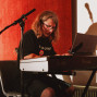 Eine Person mit langen Haaren und Brille sitzt an einem digitalen Klavier und spielt konzentriert. Um sie herum stehen Mikrofone und Musiktechnik vor einem rot beleuchteten Hintergrund.