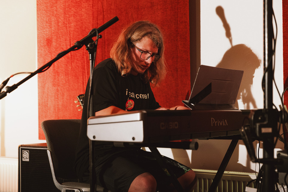 Eine Person mit langen Haaren und Brille sitzt an einem digitalen Klavier und spielt konzentriert. Um sie herum stehen Mikrofone und Musiktechnik vor einem rot beleuchteten Hintergrund.