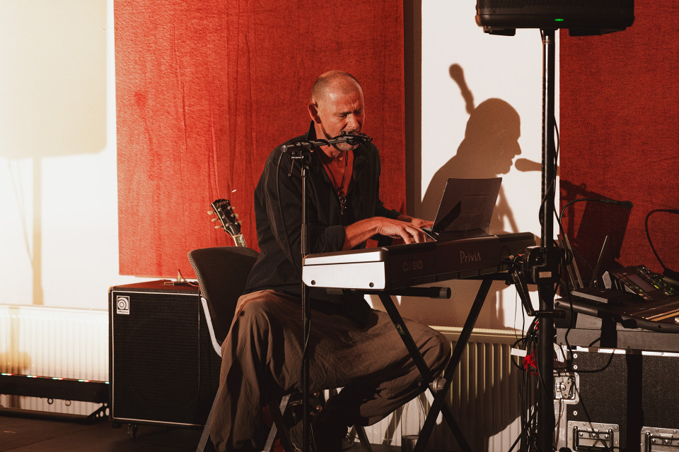 Ein Musiker sitzt auf einer kleinen Bühne am Keyboard und singt in ein Mikrofon. Warmes Licht wirft seinen Schatten auf die Wand, während Verstärker, Lautsprecher und weiteres Bühnenequipment um ihn herum stehen.
