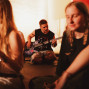 In einem warm beleuchteten Raum sitzt ein Mann auf dem Boden und spielt Bassgitarre, während um ihn herum mehrere Personen entspannt zuhören oder musizieren. Der Vordergrund ist unscharf und zeigt junge Menschen in lockerer Atmosphäre, wodurch der Fokus auf dem Musiker in der Mitte liegt.