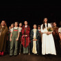 Eine Gruppe von Kindern steht auf einer dunklen Theaterbühne in verschiedenen Kostümen, darunter mittelalterliche Gewänder, Overalls und Mützen. Im Vordergrund hält ein Mädchen in einem weißen Kleid einen Blumenstrauß, während die anderen wie nach einer Aufführung posieren.