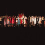 Eine Gruppe von Schauspielerinnen und Schauspielern in bunten Kostümen steht auf einer Theaterbühne und hält sich zum Schlussapplaus an den Händen. Im Vordergrund sind die dunklen Silhouetten des Publikums zu sehen, während die Bühne warm beleuchtet ist.