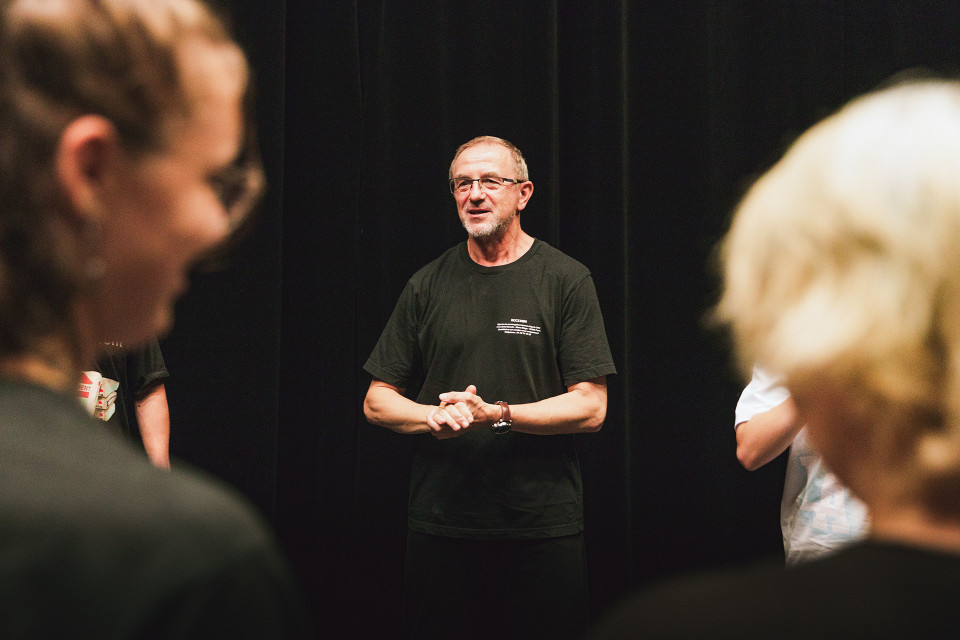 Ein Mann in einem schwarzen T-Shirt steht vor einem dunklen Vorhang und spricht zu einer Gruppe von Menschen. Im Vordergrund sind unscharf zwei Personen zu sehen, die den Blick auf die Szene wie einen Rahmen einfassen.