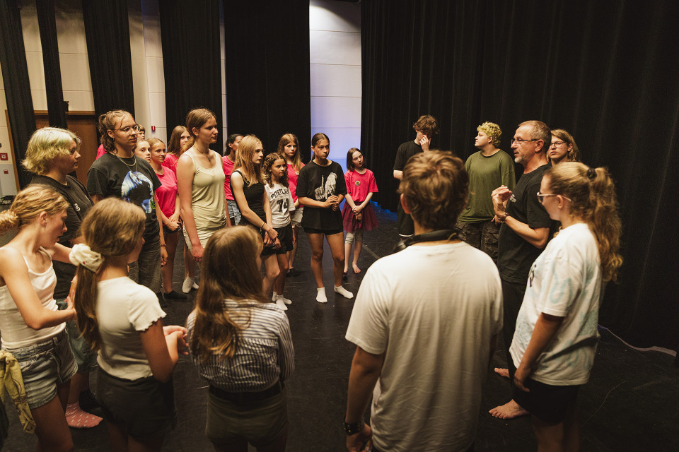 Eine Gruppe von Jugendlichen und Erwachsenen steht im Kreis auf einer Bühne vor schwarzen Vorhängen. Ein Mann spricht zur Gruppe, die aufmerksam zuhört, als ob sie an einer Theaterprobe oder einem Workshop teilnimmt.