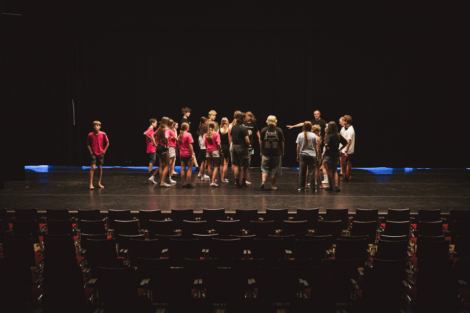 Eine Gruppe von Jugendlichen und einigen Erwachsenen steht auf einer dunklen Theaterbühne und scheint zu proben oder Anweisungen zu erhalten. Im Vordergrund sind leere Zuschauerreihen zu sehen, während die Bühne nur schwach beleuchtet ist.