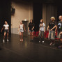 Eine Gruppe junger Menschen steht in einer Reihe auf einer Theaterbühne und scheint zu proben. Im Hintergrund sind rote Zuschauersitze, schwarze Vorhänge und gedämpftes Bühnenlicht zu sehen.