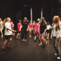 Auf einer Theaterbühne bewegen sich viele Jugendliche und ein erwachsener Mann zwischen schwarzen Vorhängen, offenbar bei einer Probe oder einem Workshop. Einige tragen pinke Shirts, andere Freizeitkleidung, und die Szene wirkt lebendig und ungezwungen.
