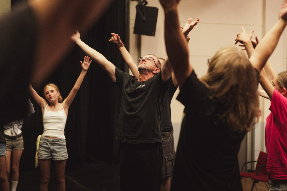 Eine Gruppe von Menschen steht in einem Raum, vermutlich bei einer Theater- oder Bewegungsprobe, und streckt die Arme nach oben. In der Mitte leitet ein Mann mit Brille die Übung, während die Teilnehmenden seine Bewegungen nachahmen.