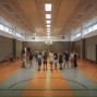 In einer großen Sporthalle steht eine Gruppe von Kindern und Jugendlichen im Kreis und hört einer Betreuerin zu. An den Wänden sind Bänke, hohe Fenster und ein Basketballkorb zu sehen.