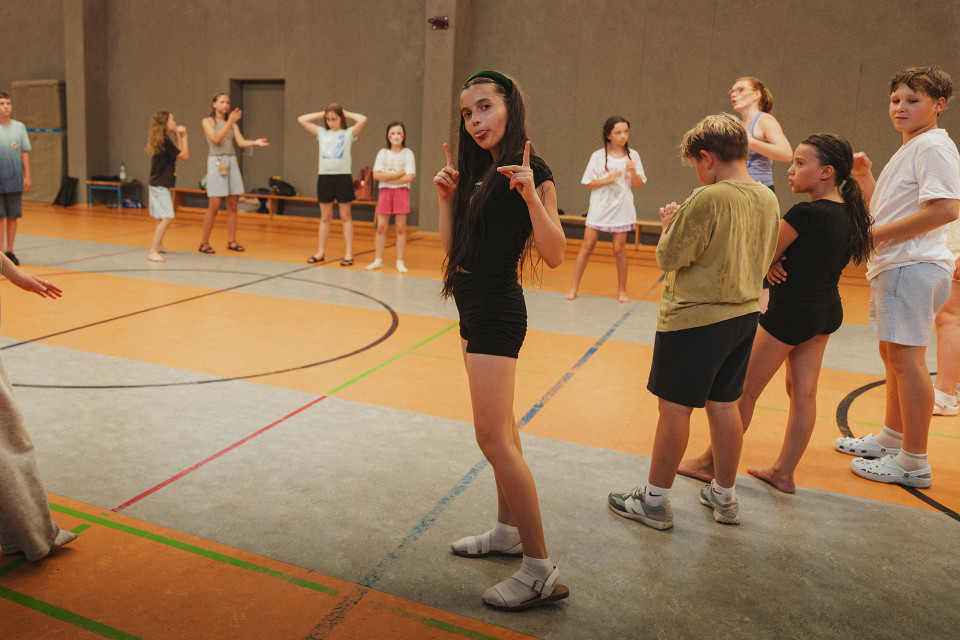 In einer Sporthalle stehen mehrere Kinder und Jugendliche verteilt auf dem Spielfeld, einige barfuß oder in Sportsachen. Im Vordergrund posiert ein Mädchen in schwarzer Kleidung mit erhobenen Fingern und herausgestreckter Zunge, während die anderen sich unterhalten oder warten.
