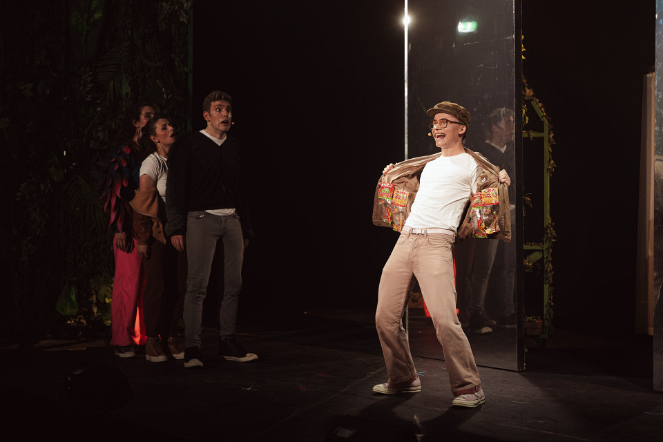 Auf einer dunklen Theaterbühne steht rechts ein hell beleuchteter Darsteller mit Kappe und Brille, der lachend seine Jacke öffnet und darin mehrere Süßigkeitentüten zeigt. Links beobachten ihn mehrere andere Darsteller aus dem Schatten heraus, während Spiegel und Bühnenkulissen die Szene rahmen.