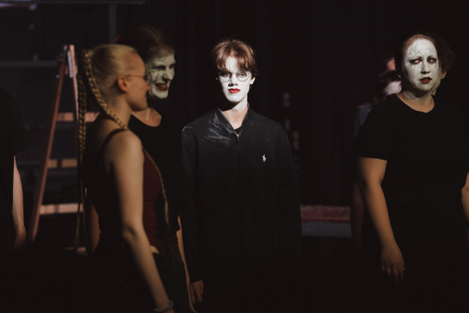 Mehrere Personen stehen in einem dunklen Raum oder auf einer Bühne, einige mit weiß geschminkten Gesichtern und dramatischem Make-up. Im Mittelpunkt steht eine Person mit Brille und schwarzer Jacke, die vom Licht angestrahlt wird, während die Umgebung weitgehend im Schatten liegt.