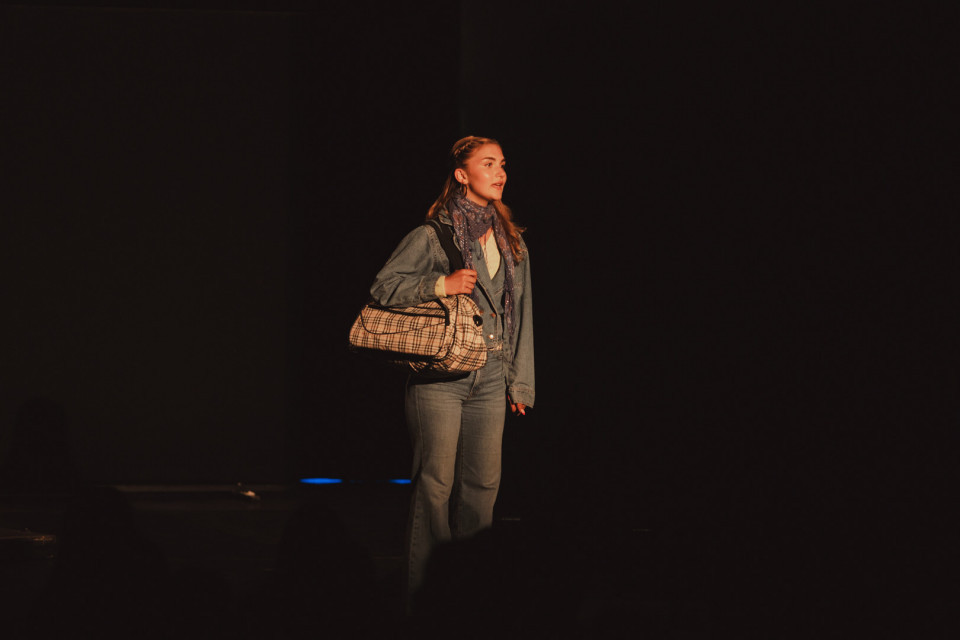 Eine junge Frau steht im Scheinwerferlicht auf einer dunklen Bühne und trägt Jeans, eine Jeansjacke sowie einen Schal. Über der Schulter hält sie eine karierte Tasche und blickt nach rechts in den Raum.