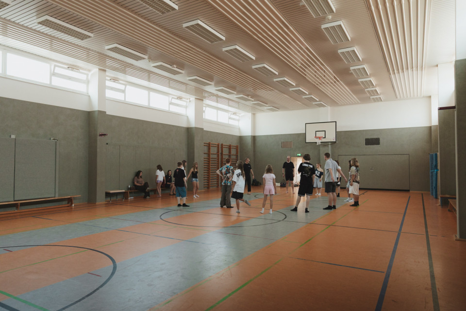 In einer großen Sporthalle stehen mehrere Jugendliche und Erwachsene auf dem Spielfeld, offenbar während eines Trainings oder Spiels. An der Wand hängt ein Basketballkorb, und durch die hohen Fenster fällt viel Licht in die Halle.