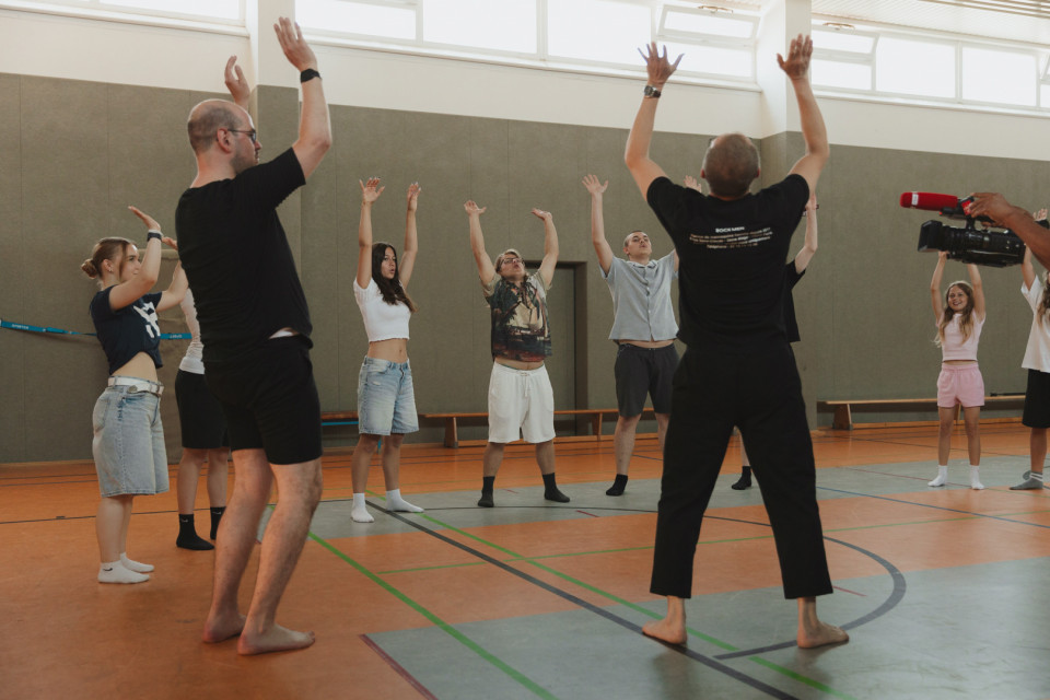 In einer Sporthalle stehen mehrere Personen im Kreis und heben gemeinsam die Arme, offenbar bei einer Übung oder einem Workshop. Rechts filmt ein Kameramann die Gruppe, während zwei Leiter im Vordergrund die Bewegung vormachen.