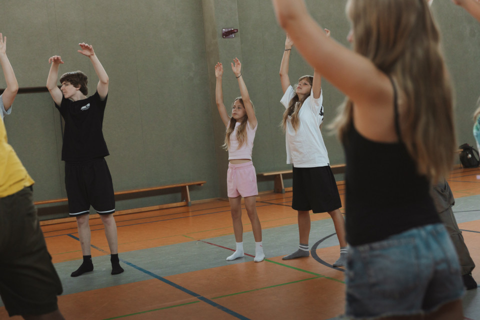 Mehrere Kinder und Jugendliche stehen in einer Sporthalle und heben bei einer Übung die Arme nach oben. Im Vordergrund ist eine Person unscharf zu sehen, während im Hintergrund Bänke und Hallenmarkierungen sichtbar sind.