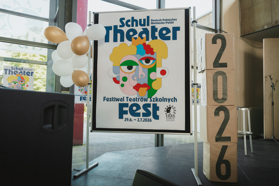 Ein großes Plakat kündigt das deutsch-polnische Schultheater-Festival „Fest“ 2026 an und zeigt eine bunte, stilisierte Gesichtsillustration. Daneben stehen weiße und goldene Luftballons sowie gestapelte Kartons mit der Jahreszahl 2026 in einem hellen Innenraum.