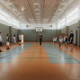 In einer hellen Sporthalle stehen mehrere Personen in zwei Reihen einander gegenüber, während eine Person in der Mitte offenbar etwas erklärt. Im Hintergrund sind ein Basketballkorb, Turnbänke und hohe Fenster zu sehen.