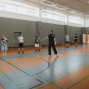 In einer großen Sporthalle stehen mehrere Jugendliche und zwei Erwachsene barfuß verteilt auf dem Spielfeld. Eine Person in der Mitte scheint die Gruppe anzuleiten oder eine Übung zu erklären.