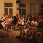 Eine Gruppe von Kindern, Jugendlichen und Erwachsenen sitzt in einem warm beleuchteten Saal auf Stühlen und schaut aufmerksam nach vorne. Links ist der Rand einer Bühne zu sehen, während im Hintergrund große Fenster und weitere Zuschauer erkennbar sind.