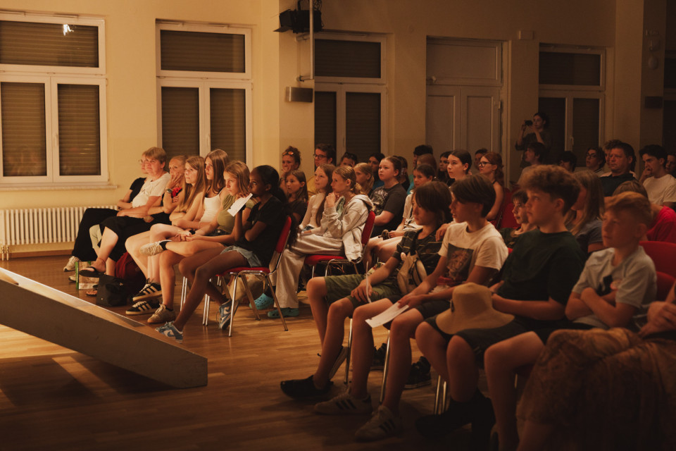 Eine Gruppe von Kindern, Jugendlichen und Erwachsenen sitzt in einem warm beleuchteten Saal auf Stühlen und schaut aufmerksam nach vorne. Links ist der Rand einer Bühne zu sehen, während im Hintergrund große Fenster und weitere Zuschauer erkennbar sind.